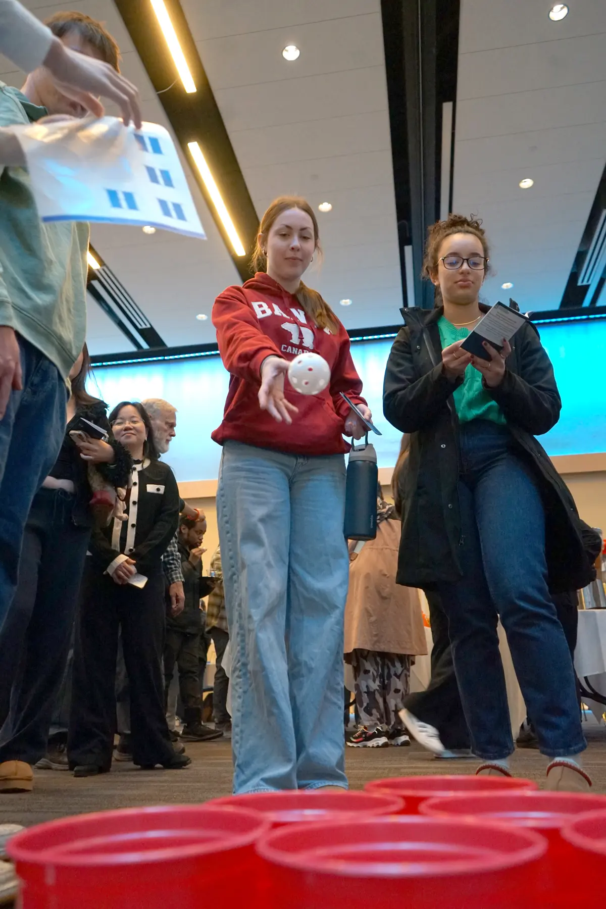 A young woman tosses a white plastic ball toward red cups while people look on in a room.