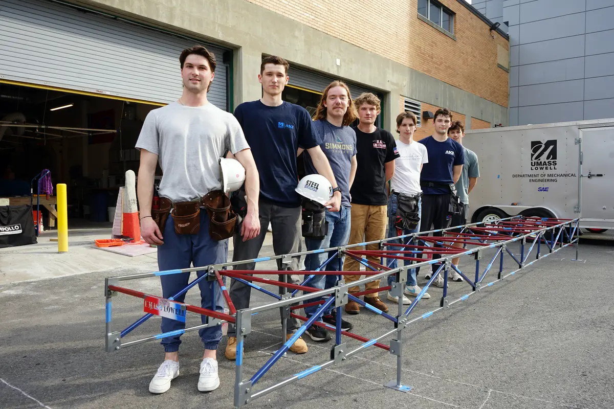Seven young men pose for a photo alongside a small steel bridge outside a garage.