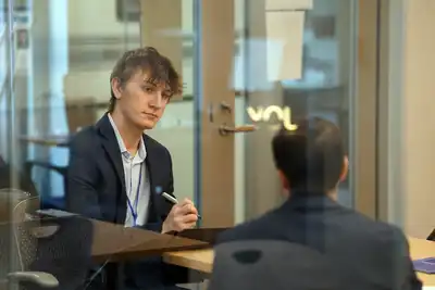 A young woman reaches across a table to shake hands with a man in a glass conference room.
