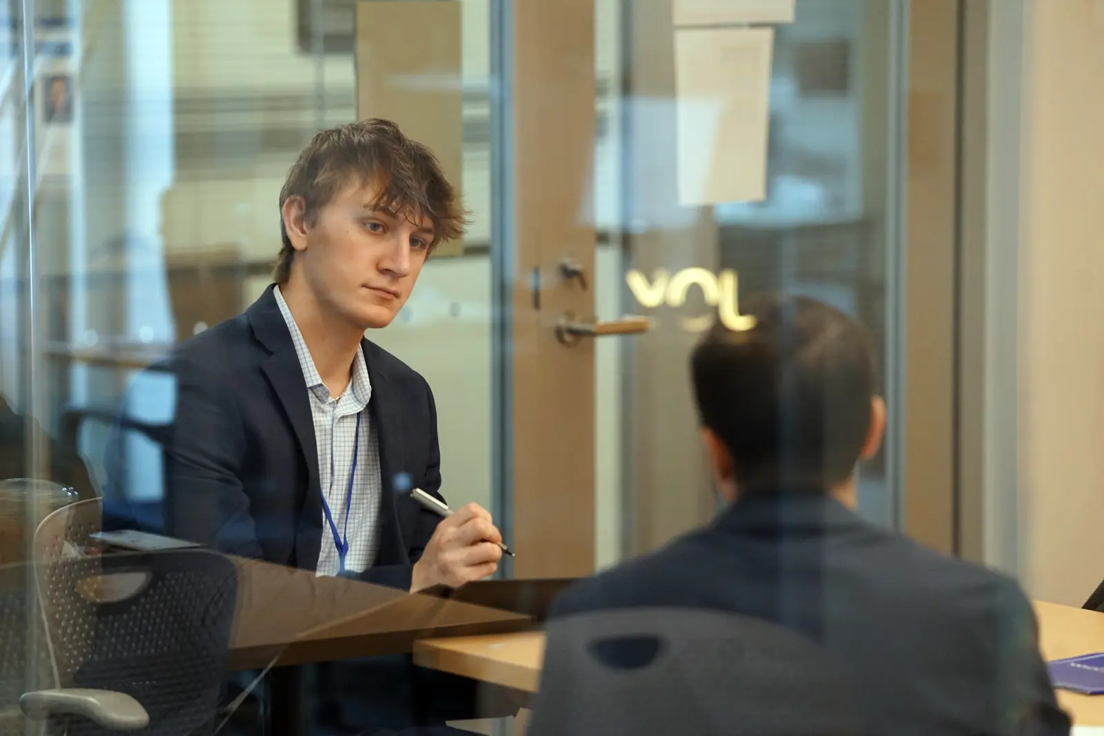 A young man listens to another person talk while holding a pen in a glass conference room.