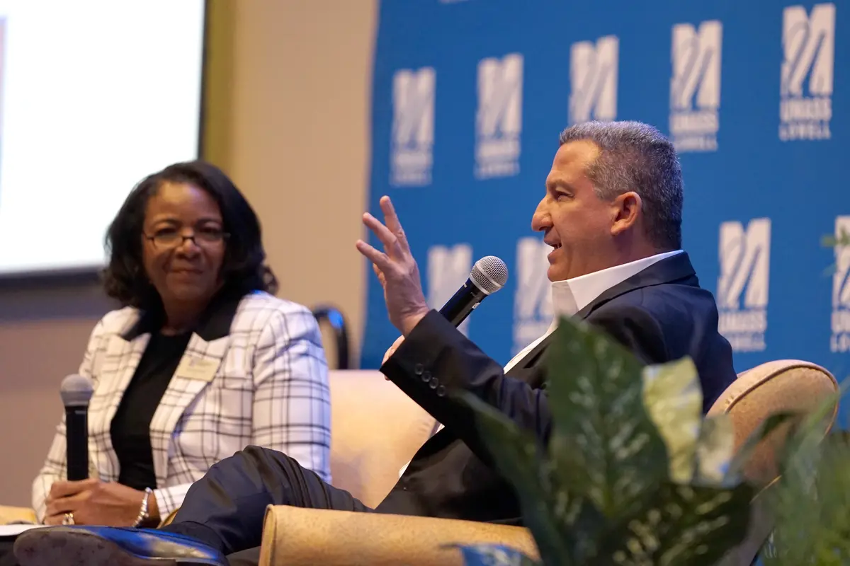 A man in a suit speaks into a microphone while sitting in a chair on a stage as a woman looks on.
