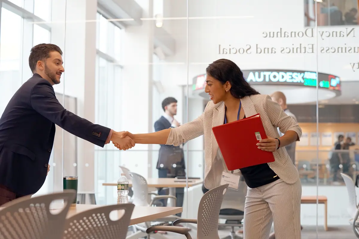 A young woman reaches across a table to shake hands with a man in a glass conference room.
