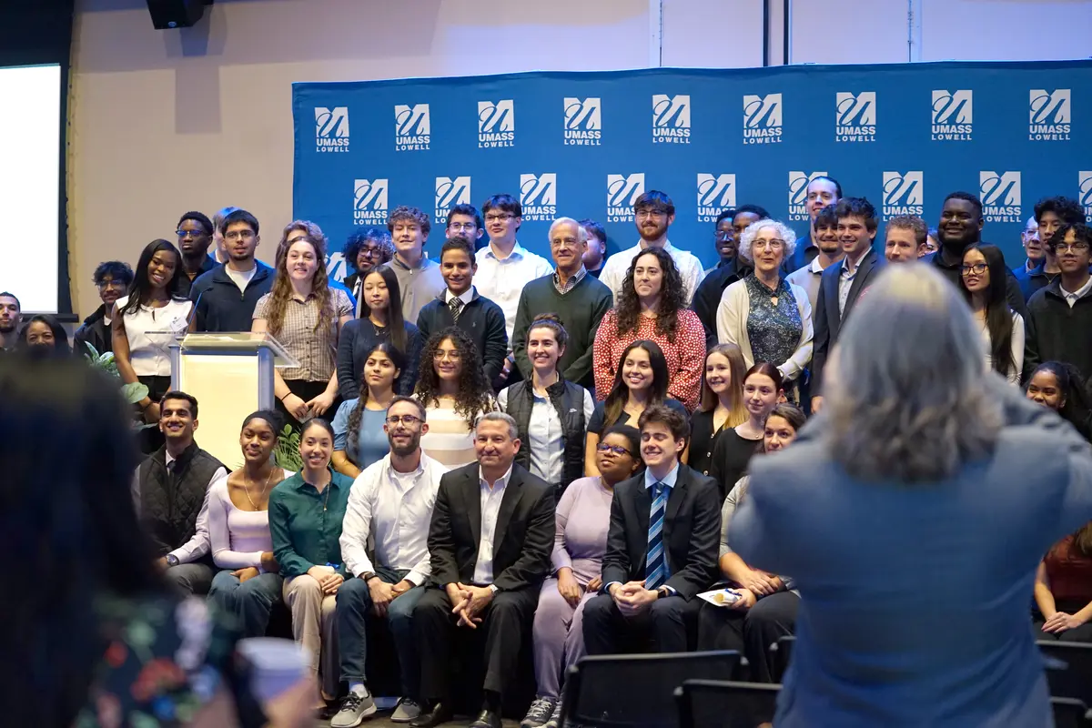 A group of about 50 people pose for a photo while on a stage in front of a blue backdrop.