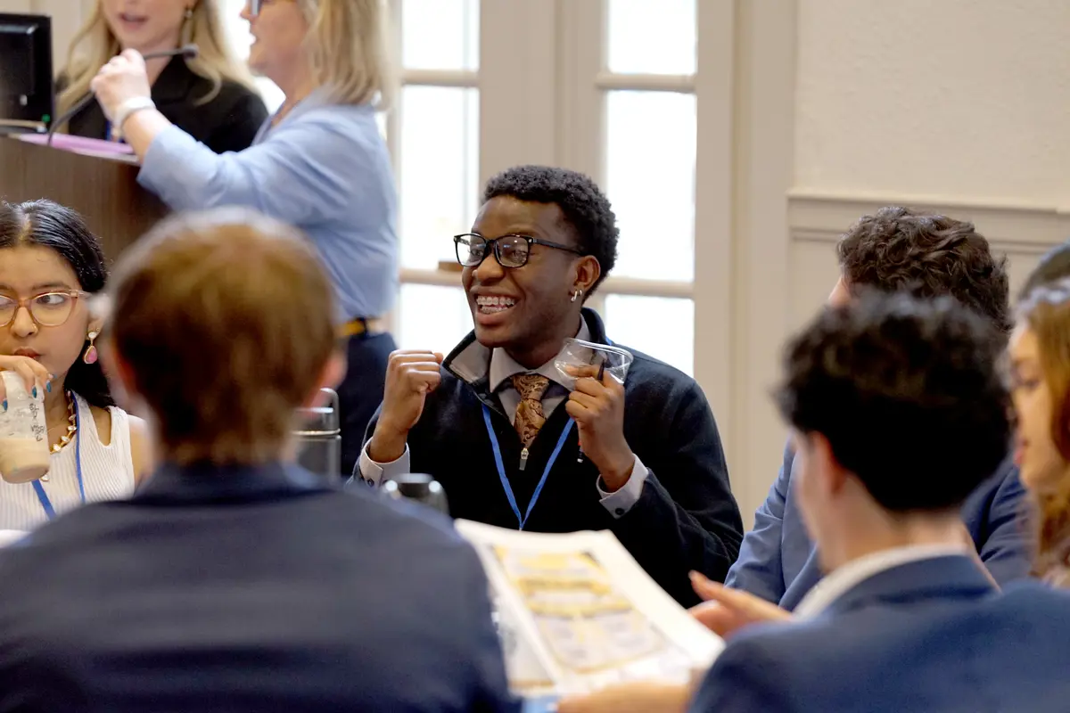A young man in glasses gives an excited reaction while seated at a table with other people.