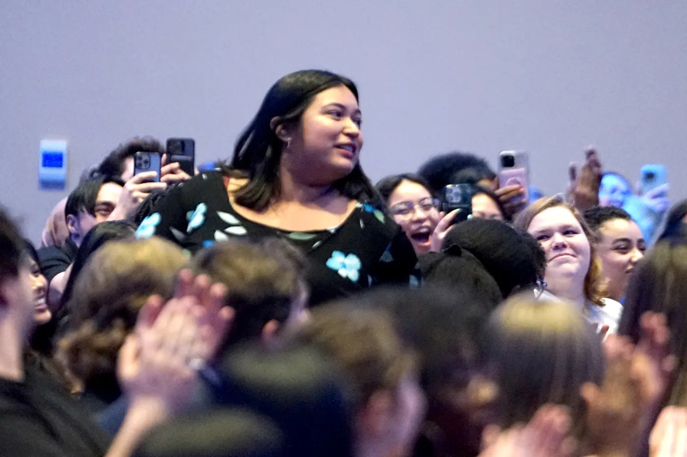 A young woman stands up in a crowd of seated students who are cheering for her.