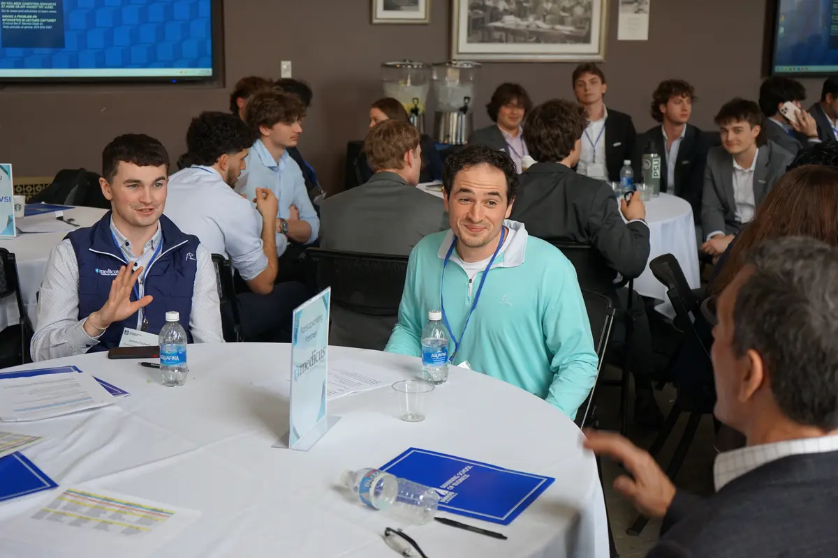 Three men talk while seated at a table in a room full of other people at tables.