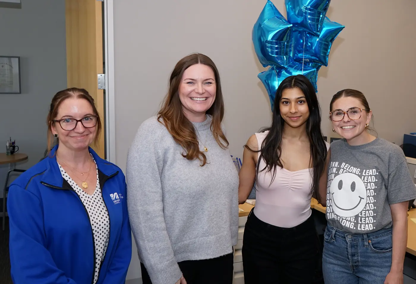 Four women pose for a photo while standing in an office. There are blue mylar balloons behind them.
