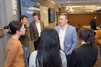 A man in a blue blazer talks to a small group of college students in a room.