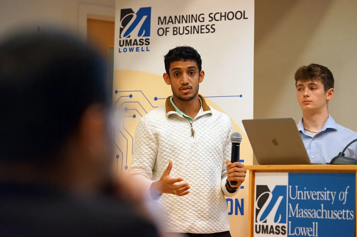 A young man holding a microphone speaks to someone while another young man looks on from a podium.
