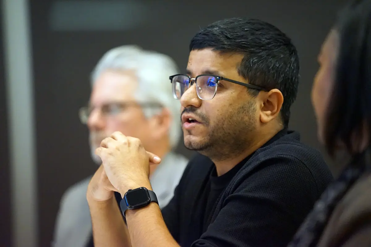 A man with dark hair and glasses speaks while seated with his hands folded in front of him.