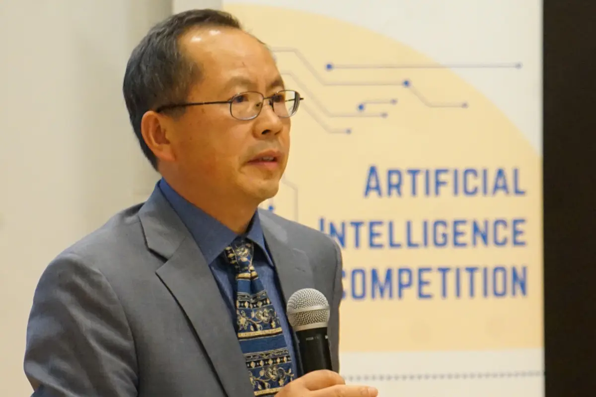 A man in a suit and glasses holds a microphone while speaking.