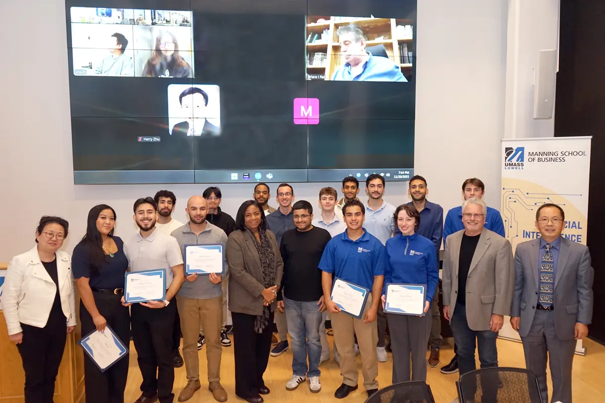 A group of 20 people pose for a group photo in front of a giant monitor on the wall behind them.