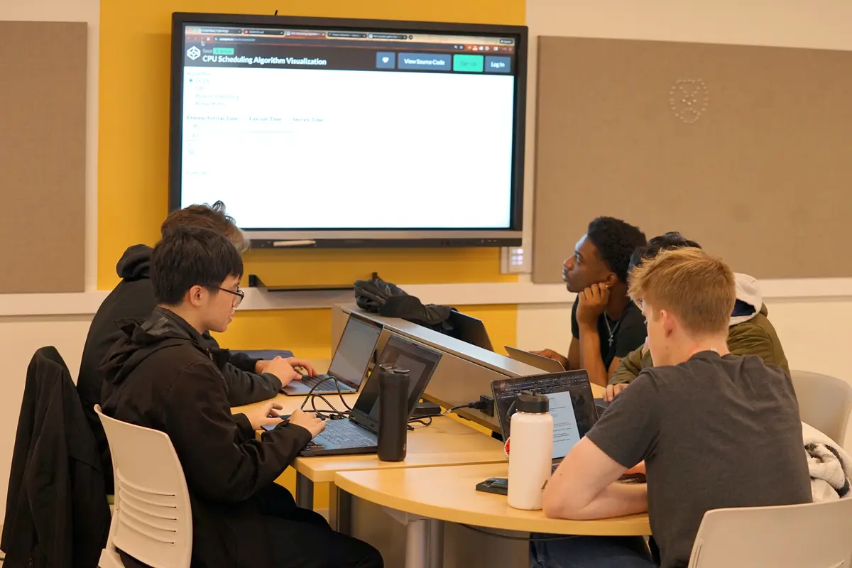 Five students sit around a table that has a monitor at the end in a classroom.
