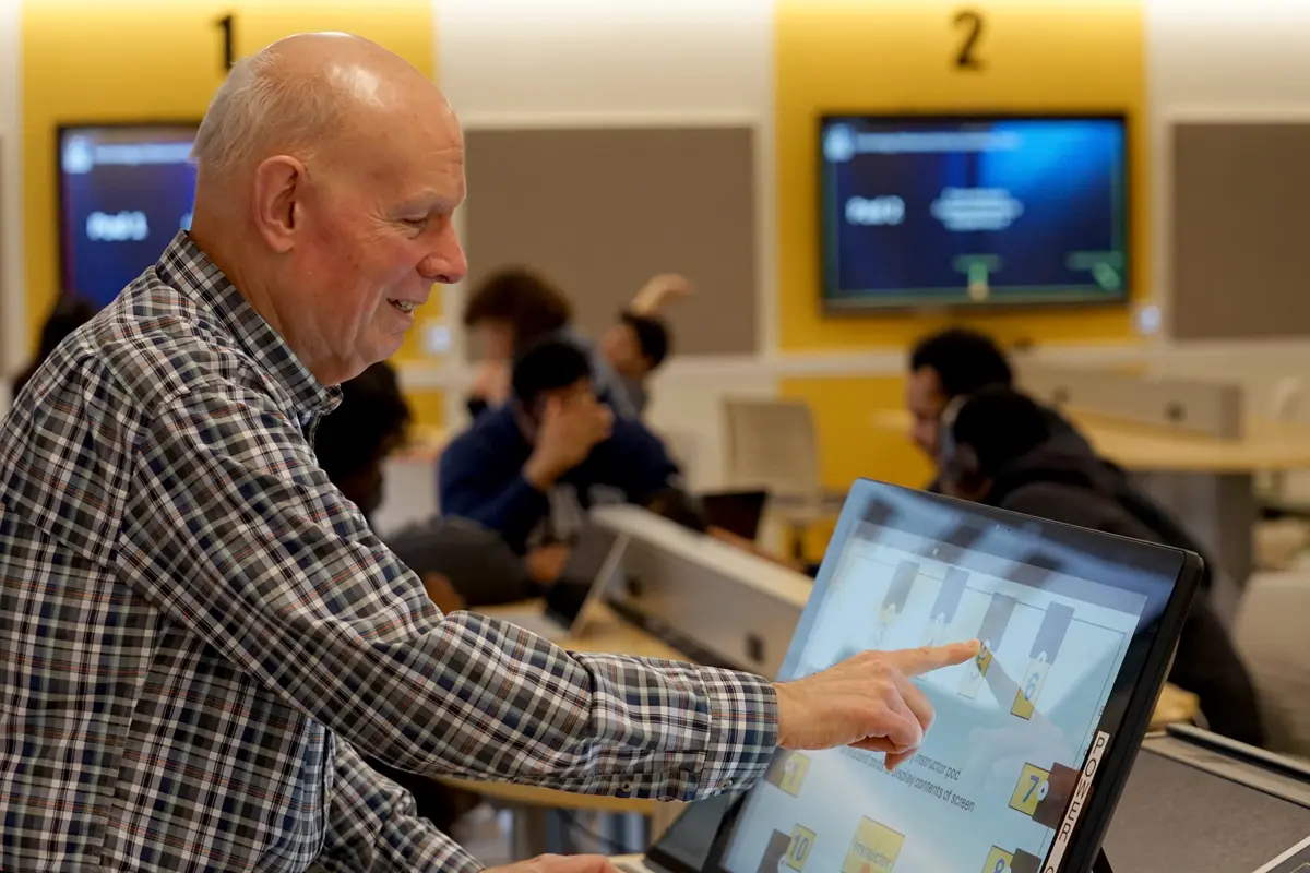 A man in a checkered shirt uses a touch screen monitor in a classroom.