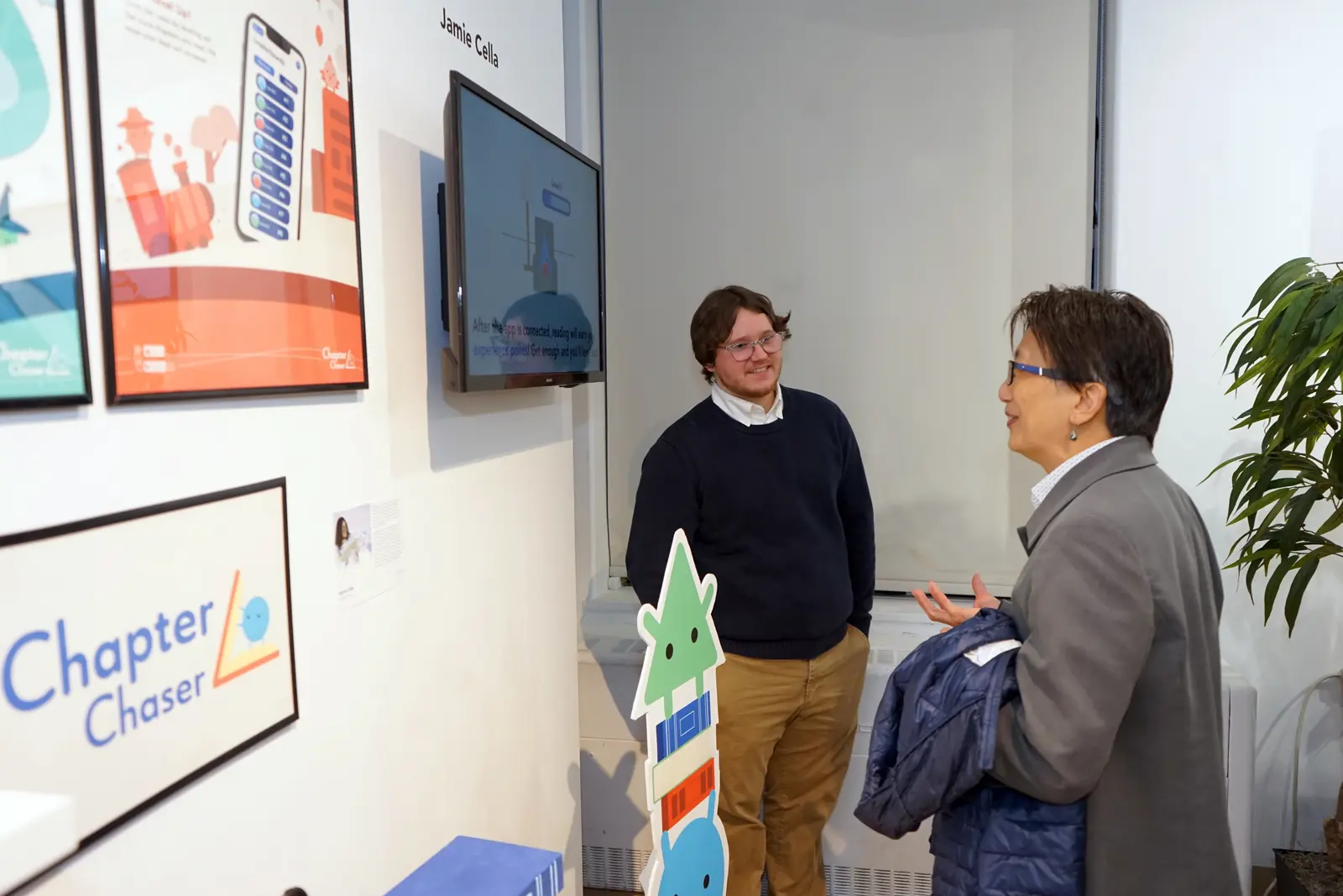 A woman with glasses looks at an art display while a young man faces her in a gallery.