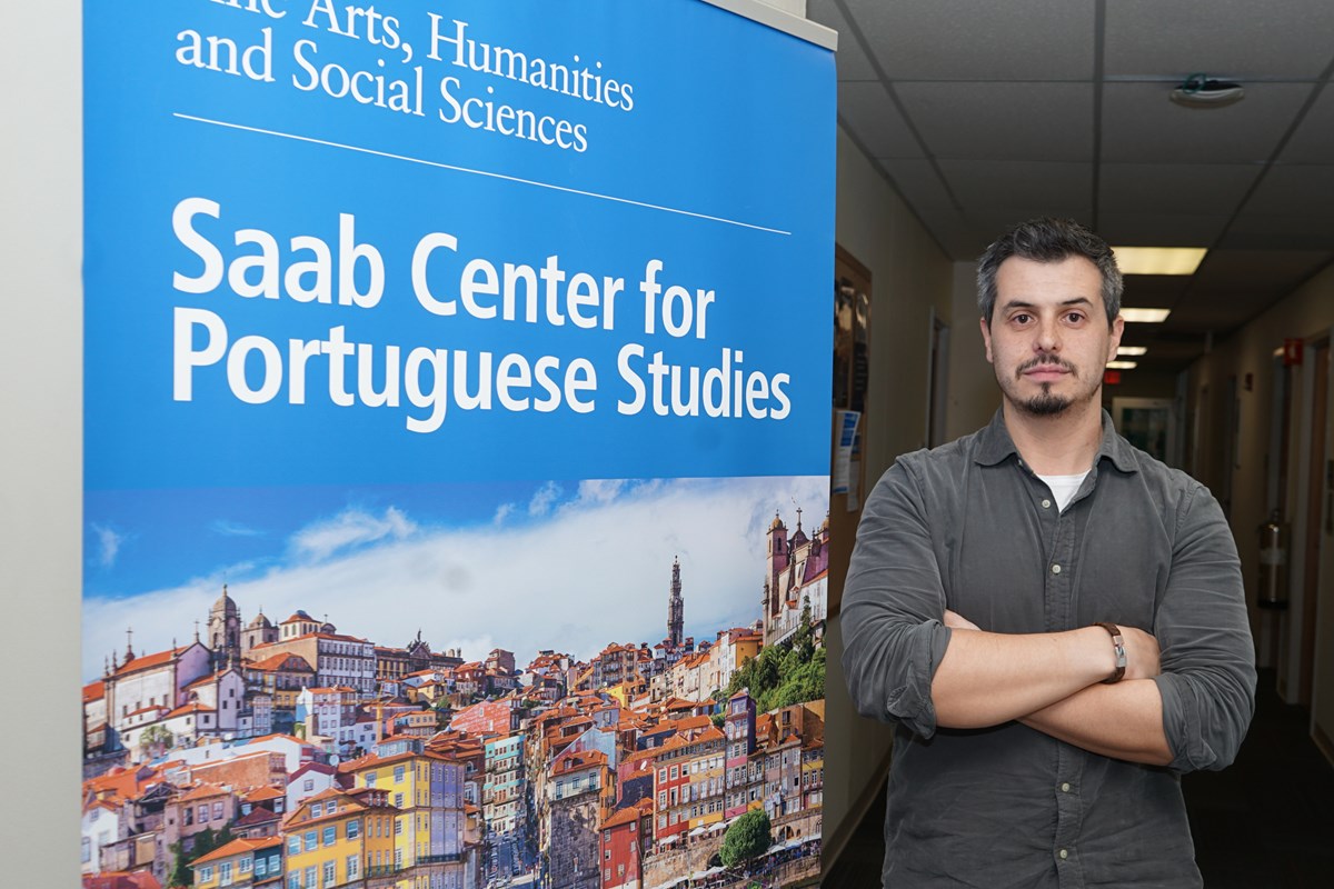 A person poses for a photo with their arms folded while standing next to a sign for the Saab Center for Portuguese Studies.