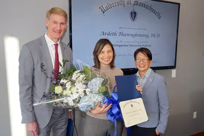 A woman holds a bouquet of flowers and a certificate while posing for a photo with a man and a woman in front of a TV screen.