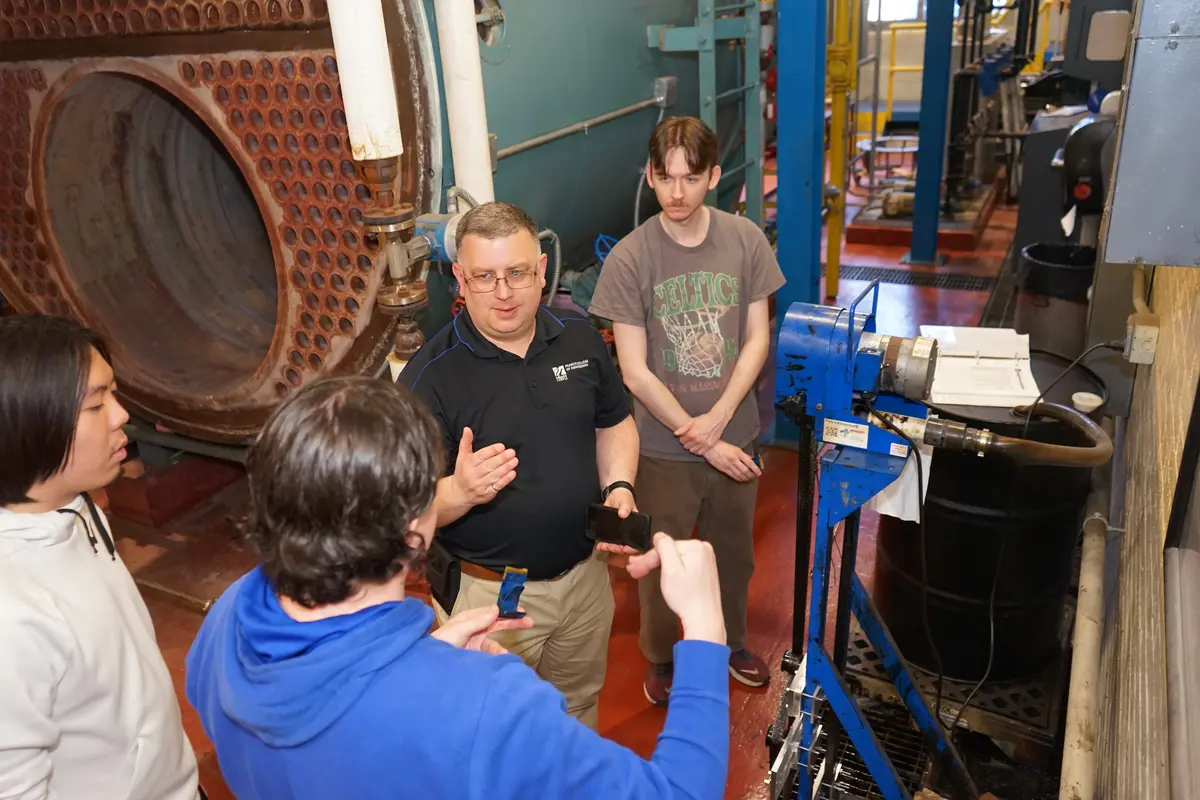 A man in glasses talks to three college students in a boiler room.
