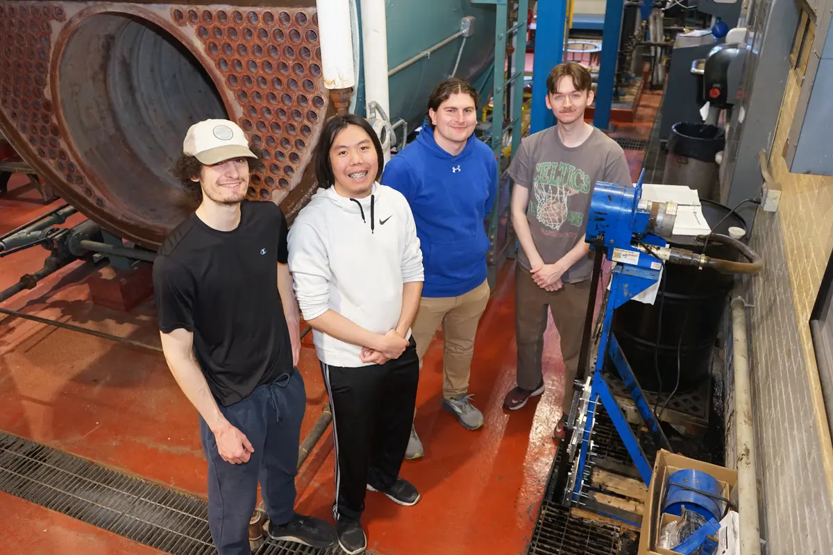 Four young men pose for a photo in front of a boiler in a power plant room.