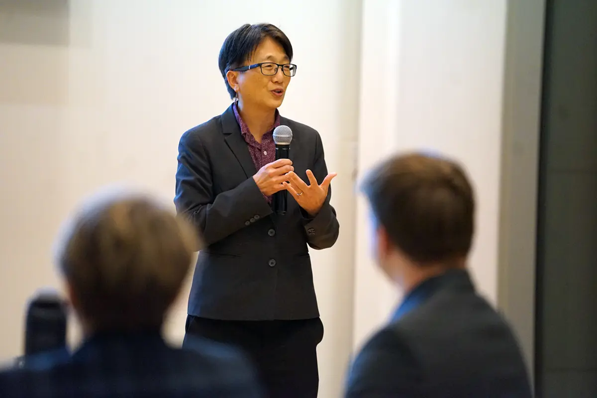 A woman in glasses and a dark blazer holds a microphone and gestures with her hand while speaking to an audience.