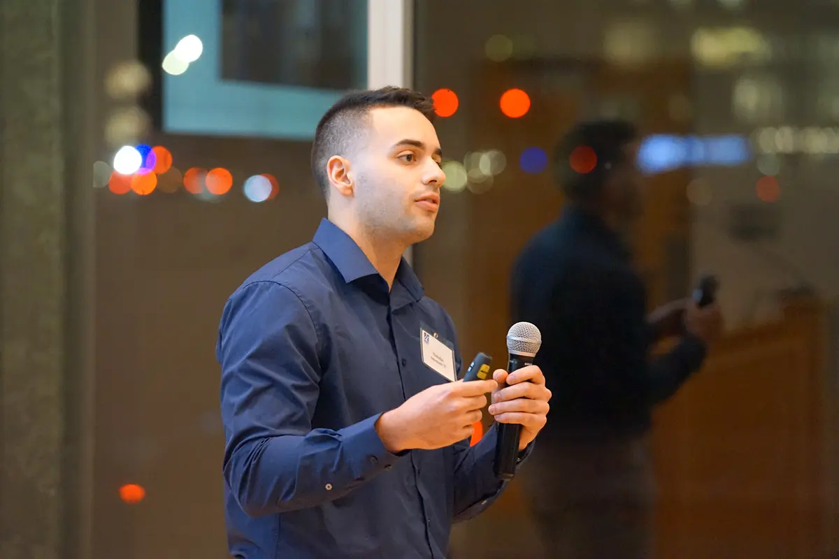 A young man in a blue shirt holds a microphone and gestures with his hand while speaking.