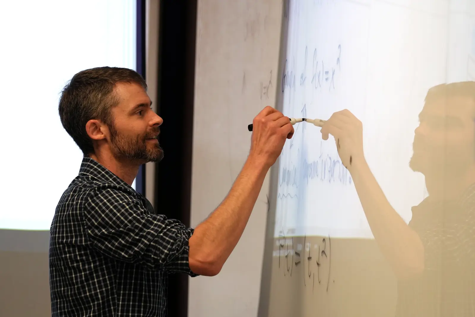 A man with his sleeves rolled up writes on a dry erase board in a classroom.