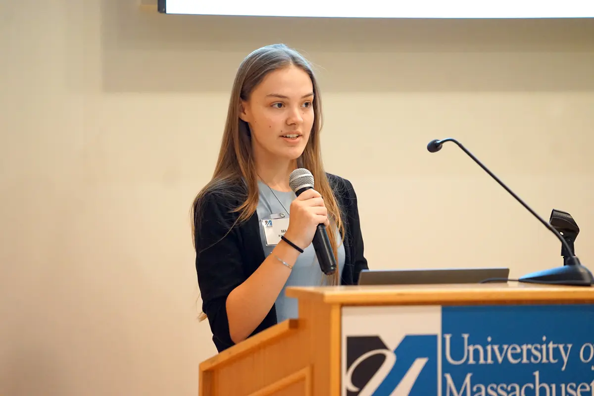 A young woman with long hair holds a microphone while speaking at a podium.