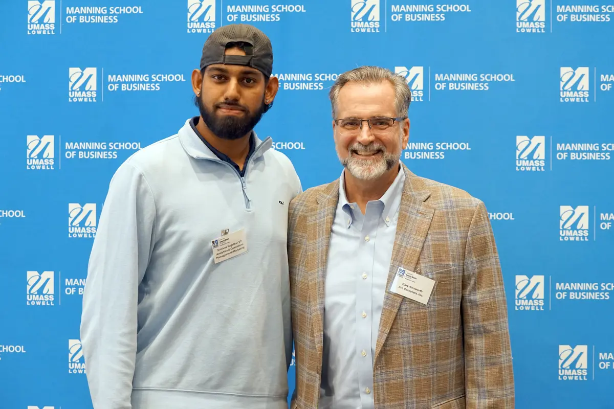 A young man wearing a backwards baseball cap poses for a photo with a man in a plaid blazer while standing in front of a blue backdrop.