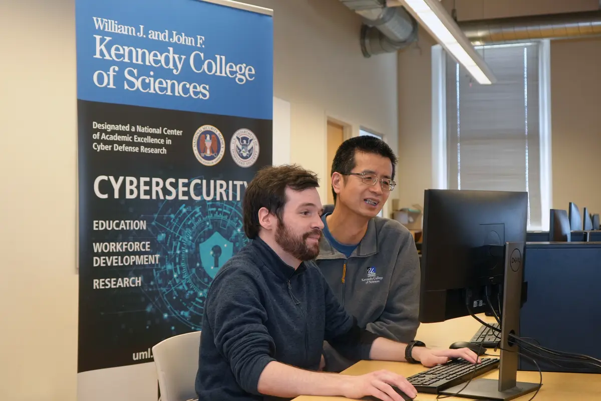 Matthew Harper and Xinwen Fu look at a computer screen with a cybersecurity sign behind them.