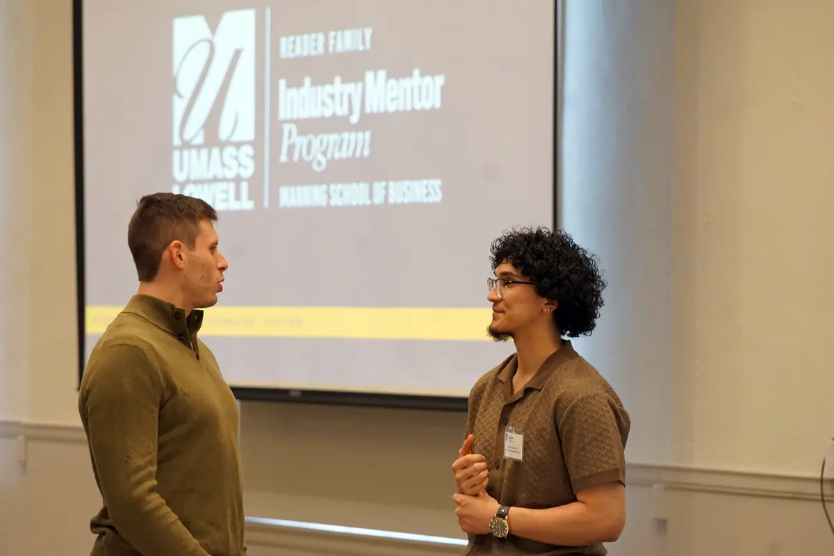 A young man with curly hair and glasses talks to a man with short hair in front of a projection screen.