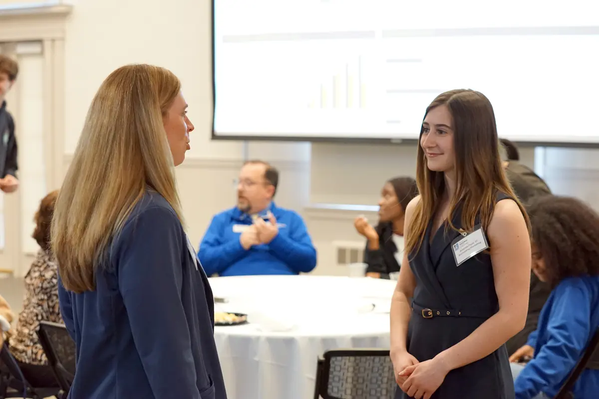 A young woman with long brown hair smiles while listening to a woman speak while standing in a room.