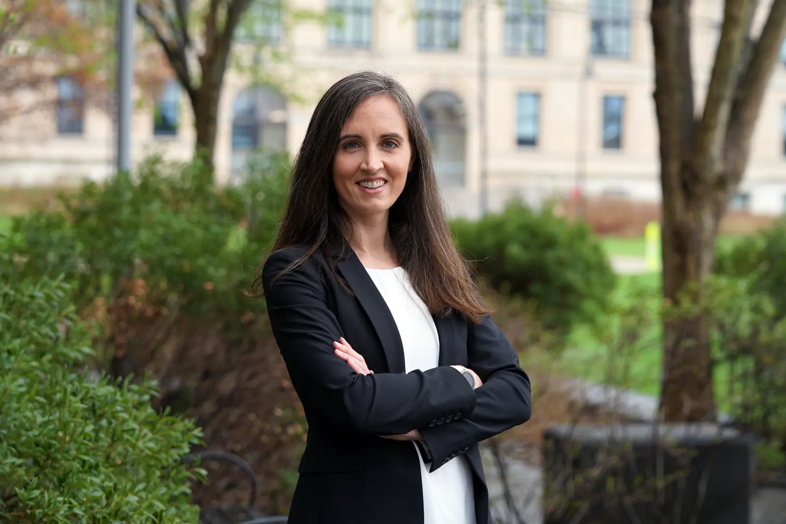 A woman poses for a photo with her arms folded while standing in front of a campus building.