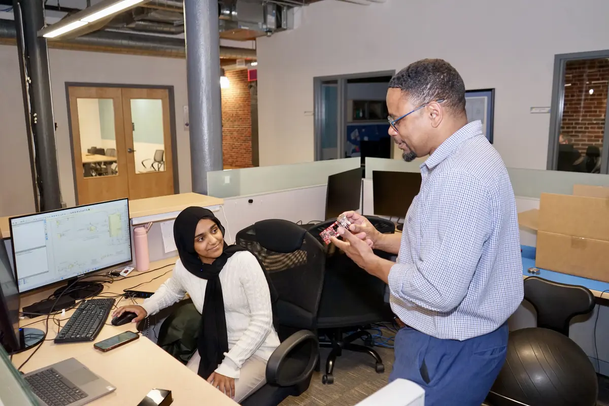 A man in glasses holds a device in his hands while talking to a young woman who is seated at a computer at a desk.