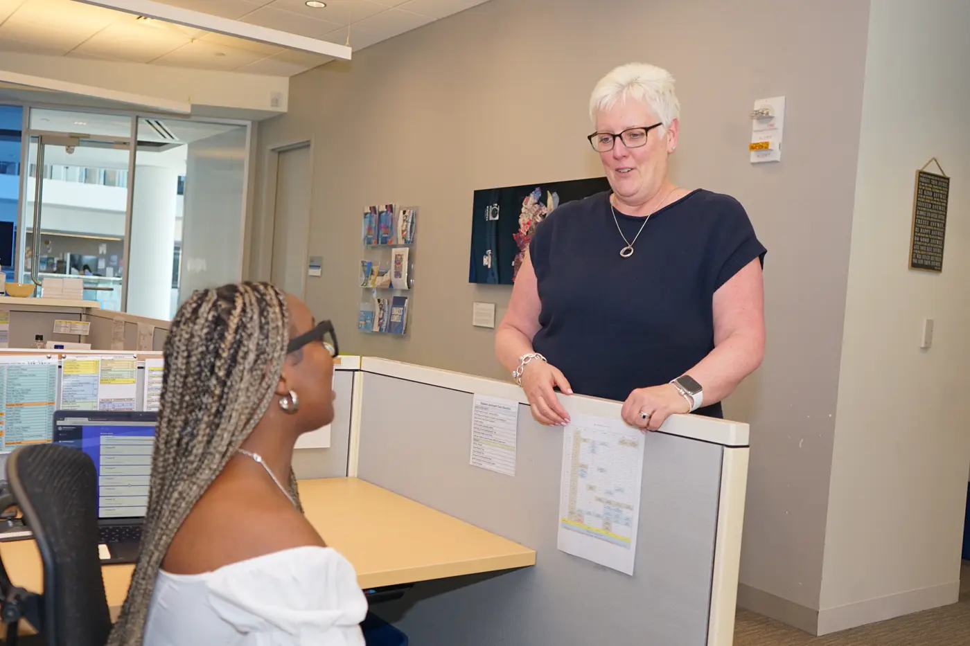 A woman with short hair and glasses stands at a cubicle and talks to a woman who is seated.