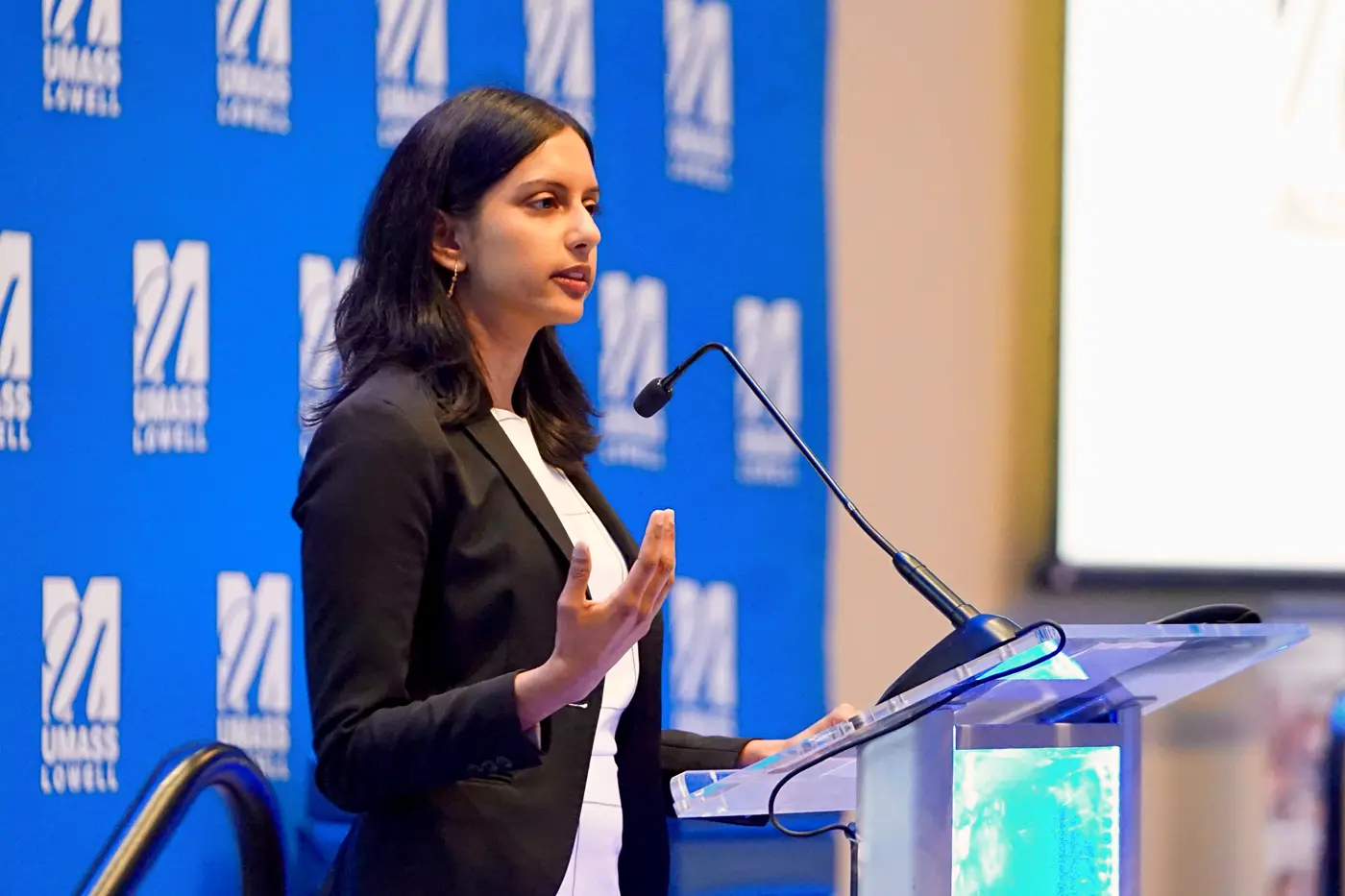 A person gestures with their hand while speaking at a podium on a stage in front of a blue backdrop.