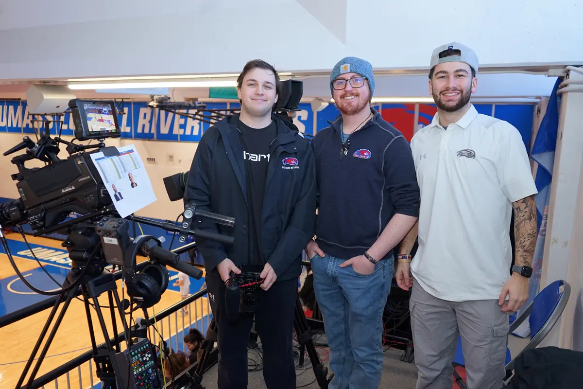 Three young men pose for a photo next to a TV camera while standing in the top row of a college basketball gym.