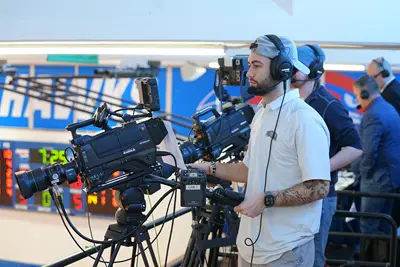 Three young men pose for a photo next to a TV camera while standing in the top row of a college basketball gym.