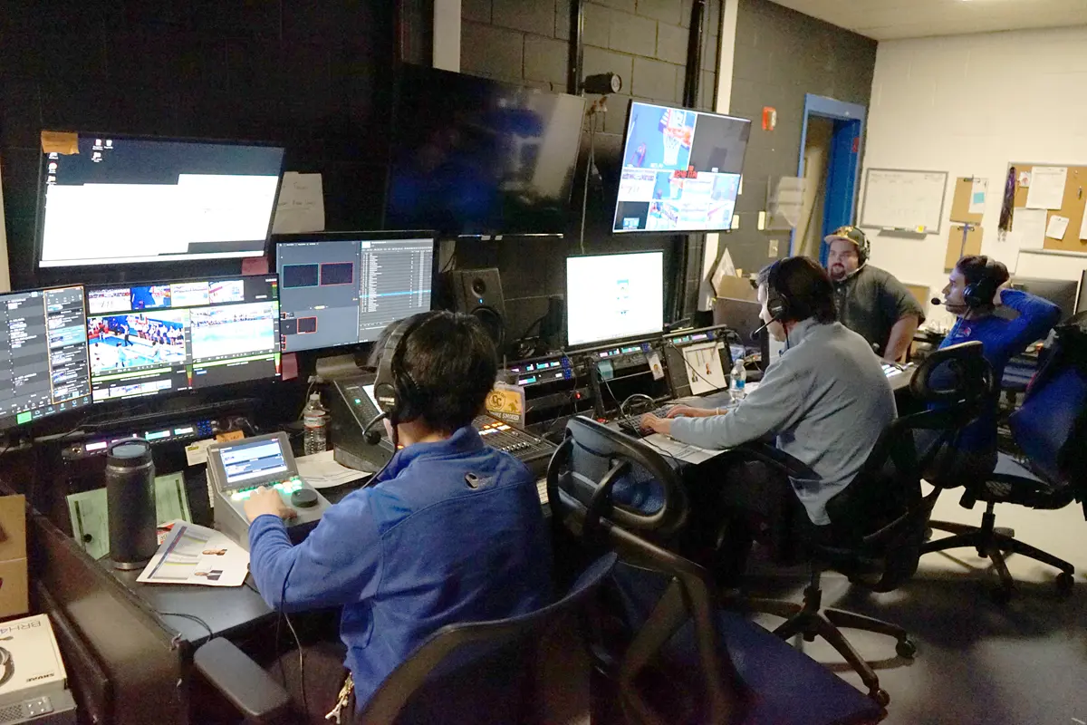 Four men sit in front of TV and computer monitors and control panels in a dark broadcast room.