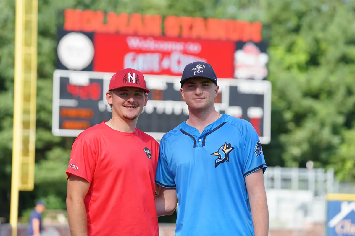 Two college baseball players, one in a red uniform and the other in blue, pose for a photo in front of a scoreboard.