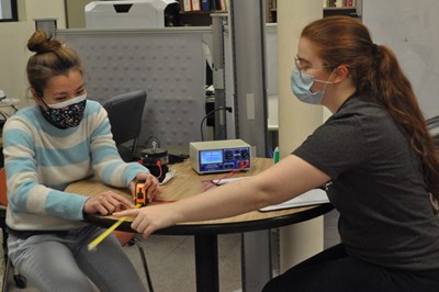 Two masked female students working in a lab using a tape measure.