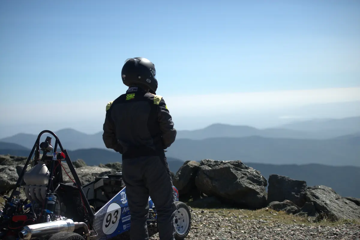 A man in a race car driving helmet stands next to a car and looks at the view from a mountain peak.