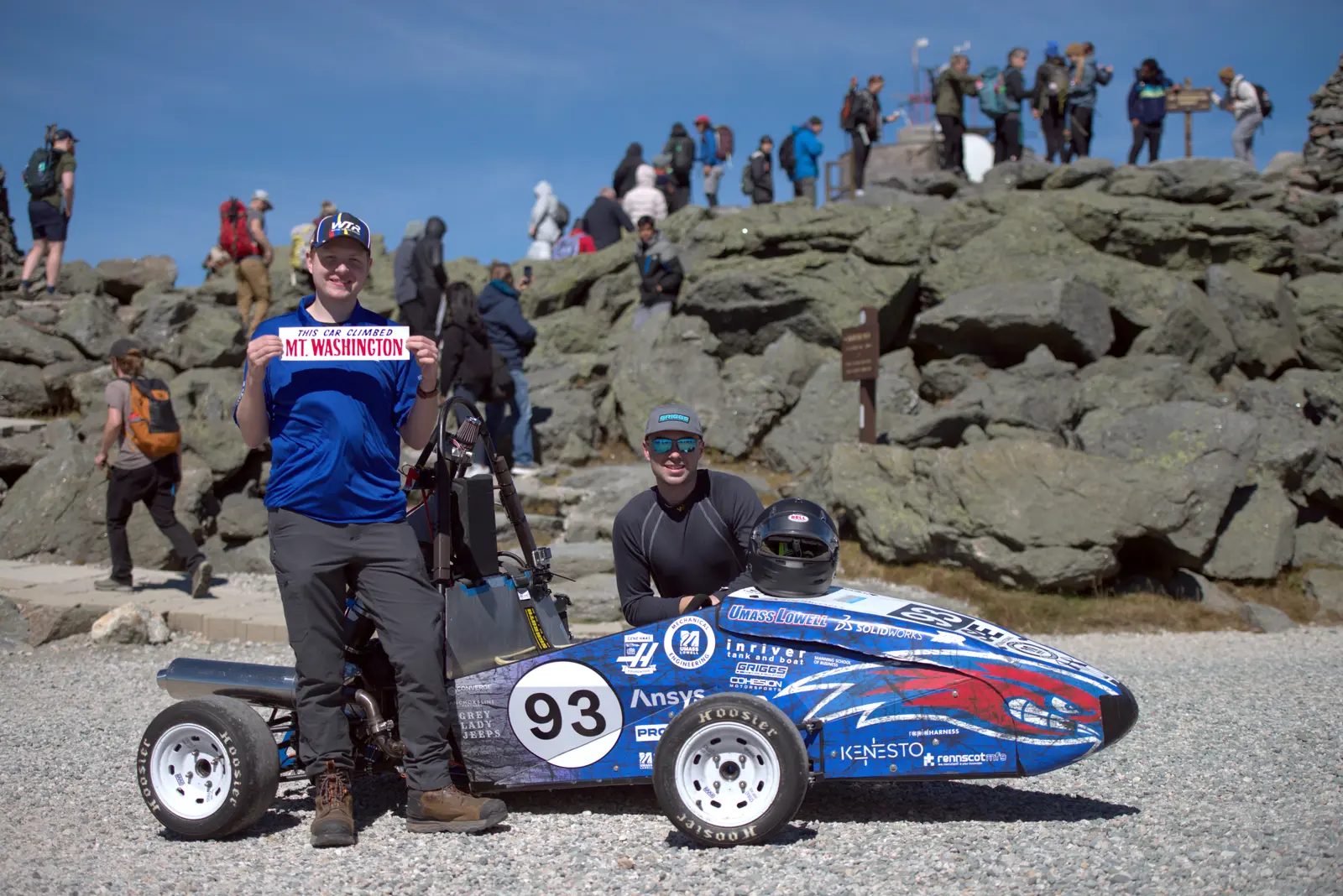 A young man holds a bumper sticker while standing next to a racecar that another man is kneeling behind. 