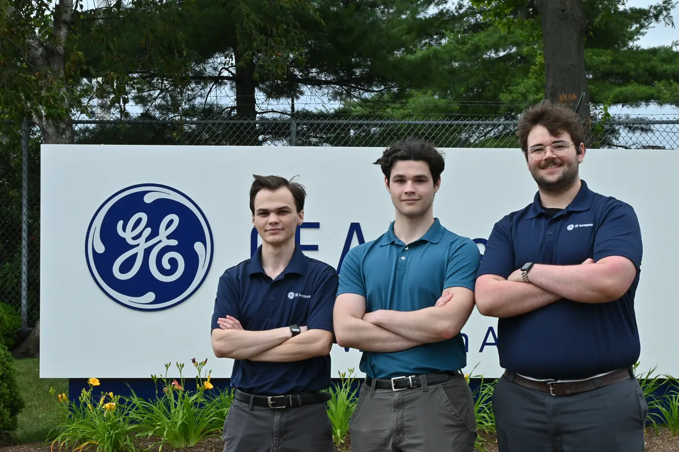 Three young men in blue polo shirts pose for a photo in front of a GE sign with their arms folded.