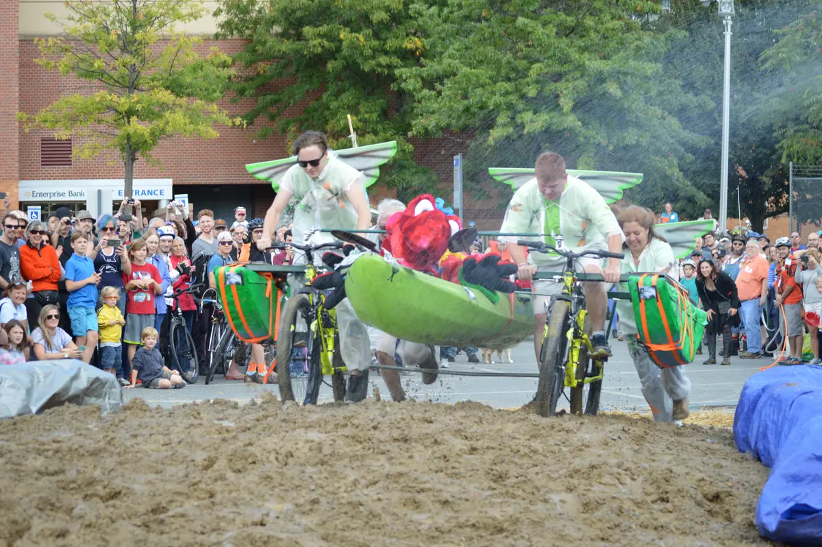 Two young men with wings on their backs pedal a bicycle sculpture through a mud pit as people cheer them on.