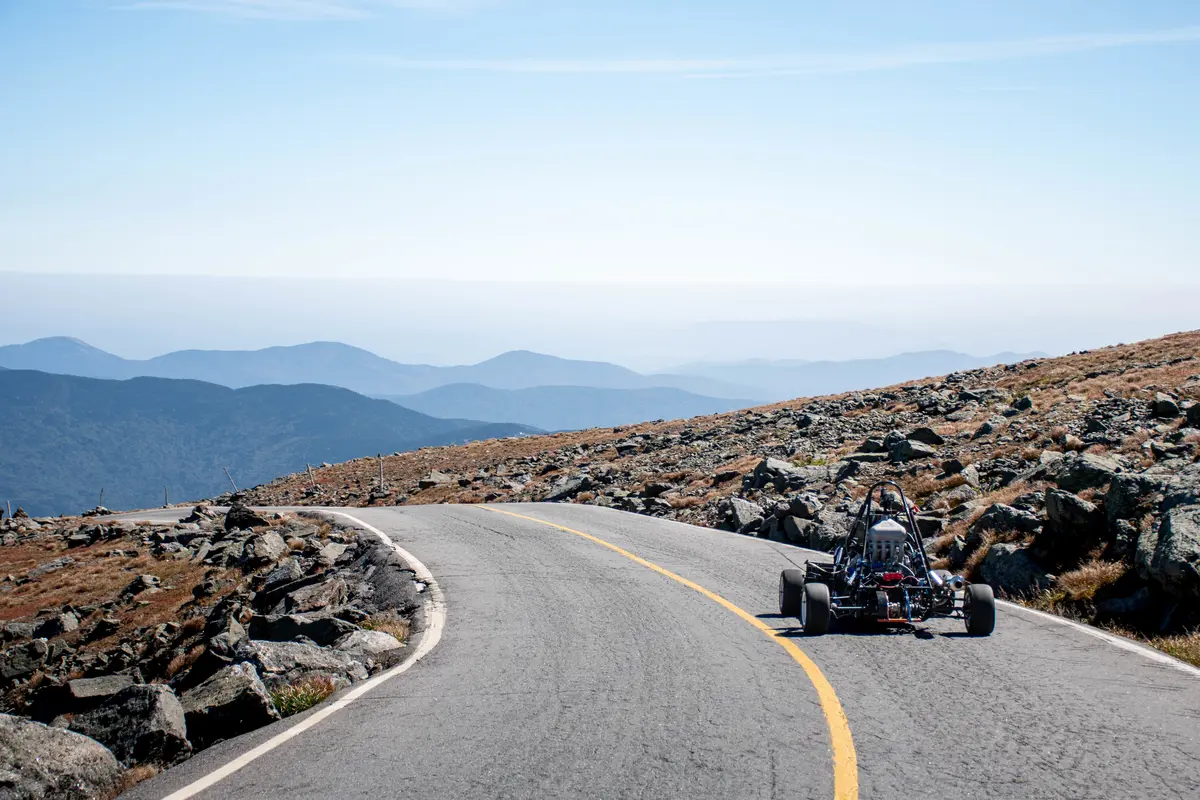 A race car drives on a mountain road, with mountains on the horizon.