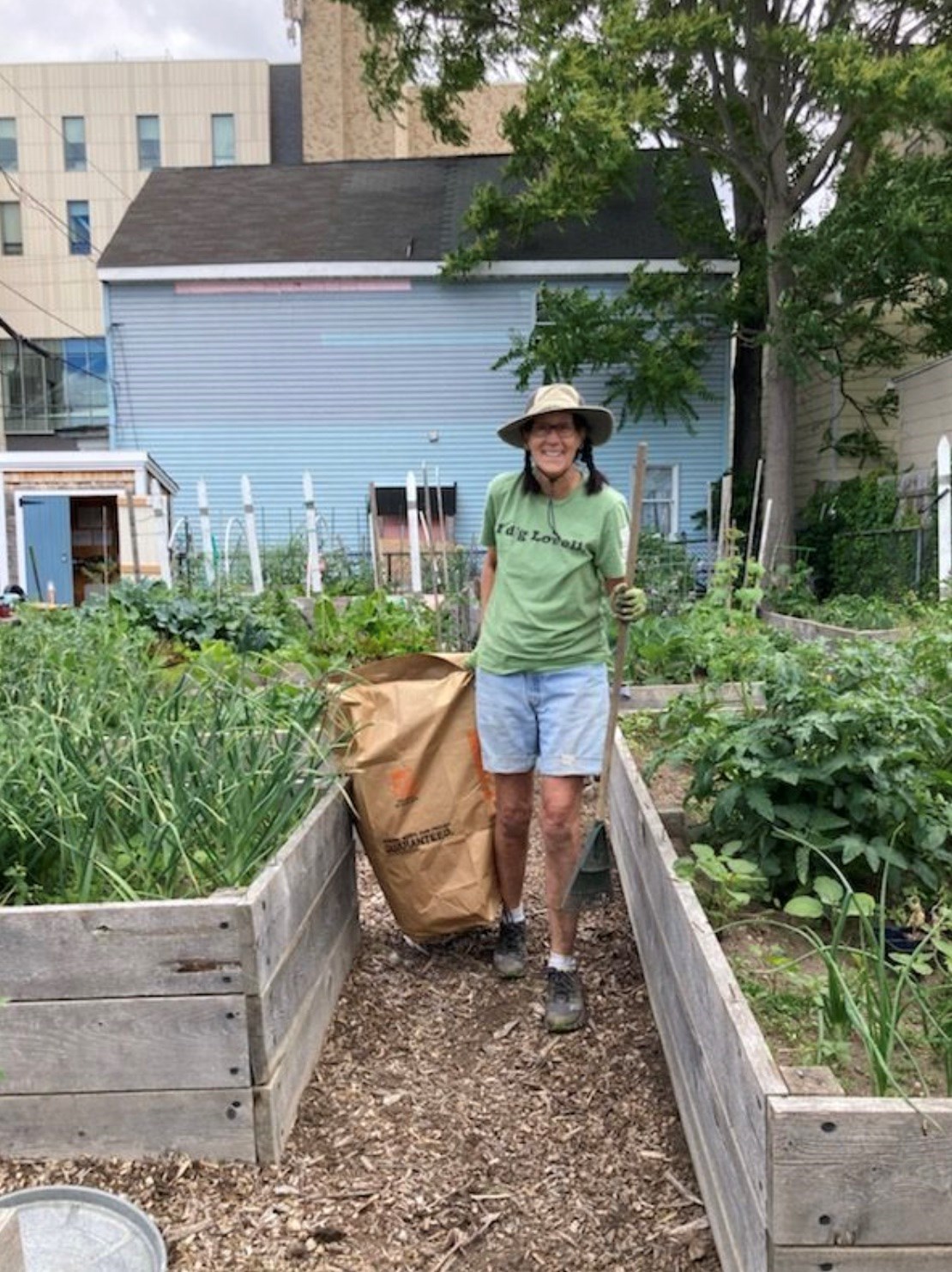 Image of woman standing in garden with rake and compost bag. The garden has several raised garden beds made from wood structures. 