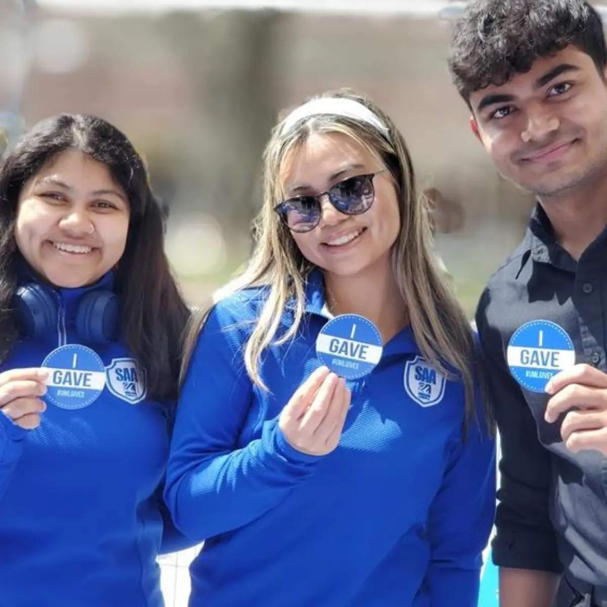 Three people wearing blue shirts hold round blue stickers that read "I GAVE" in an outdoor setting.