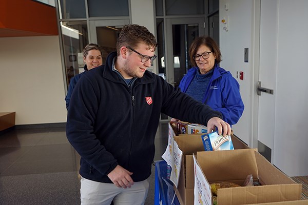 A young man in glasses puts a box of pasta in a food donation box while two women look on