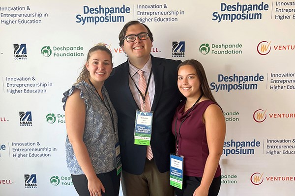 Two young women and a man in glasses pose for a photo in front of a conference backdrop