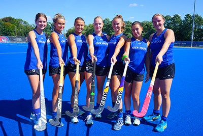 Seven college field hockey players pose for a group photo on a blue turf field